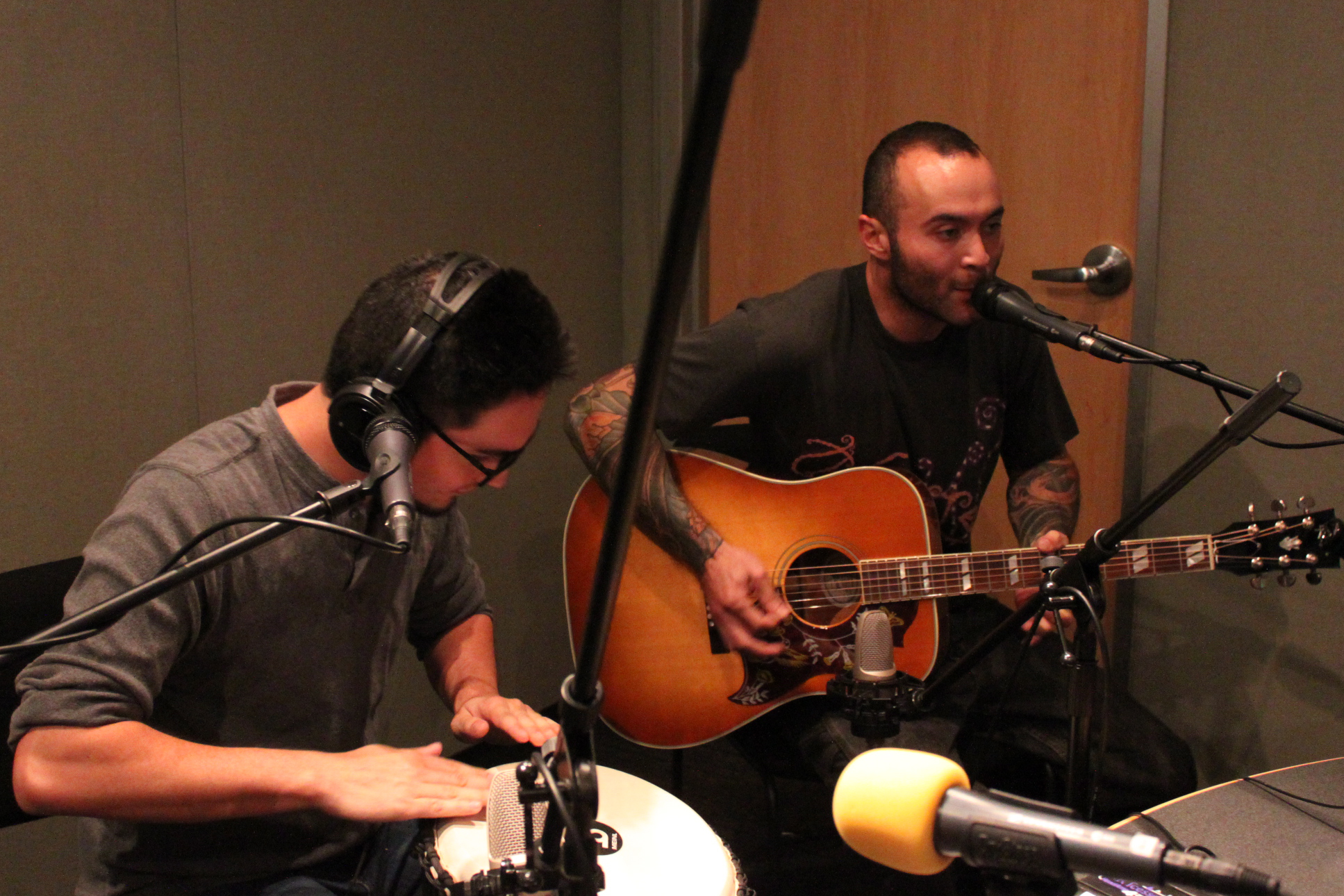 An adult wearing headphones playing a conga and an adult playing an acoustic guitar and singing into a microphone in a radio studio