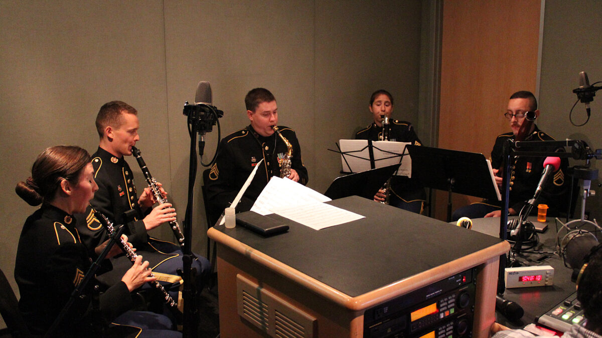 An adult playing oboe, an adult playing clarinet, an adult playing alto saxophone, an adult playing bass clarinet, and an adult playing bassoon, all looking at sheet music on music stands in a radio studio