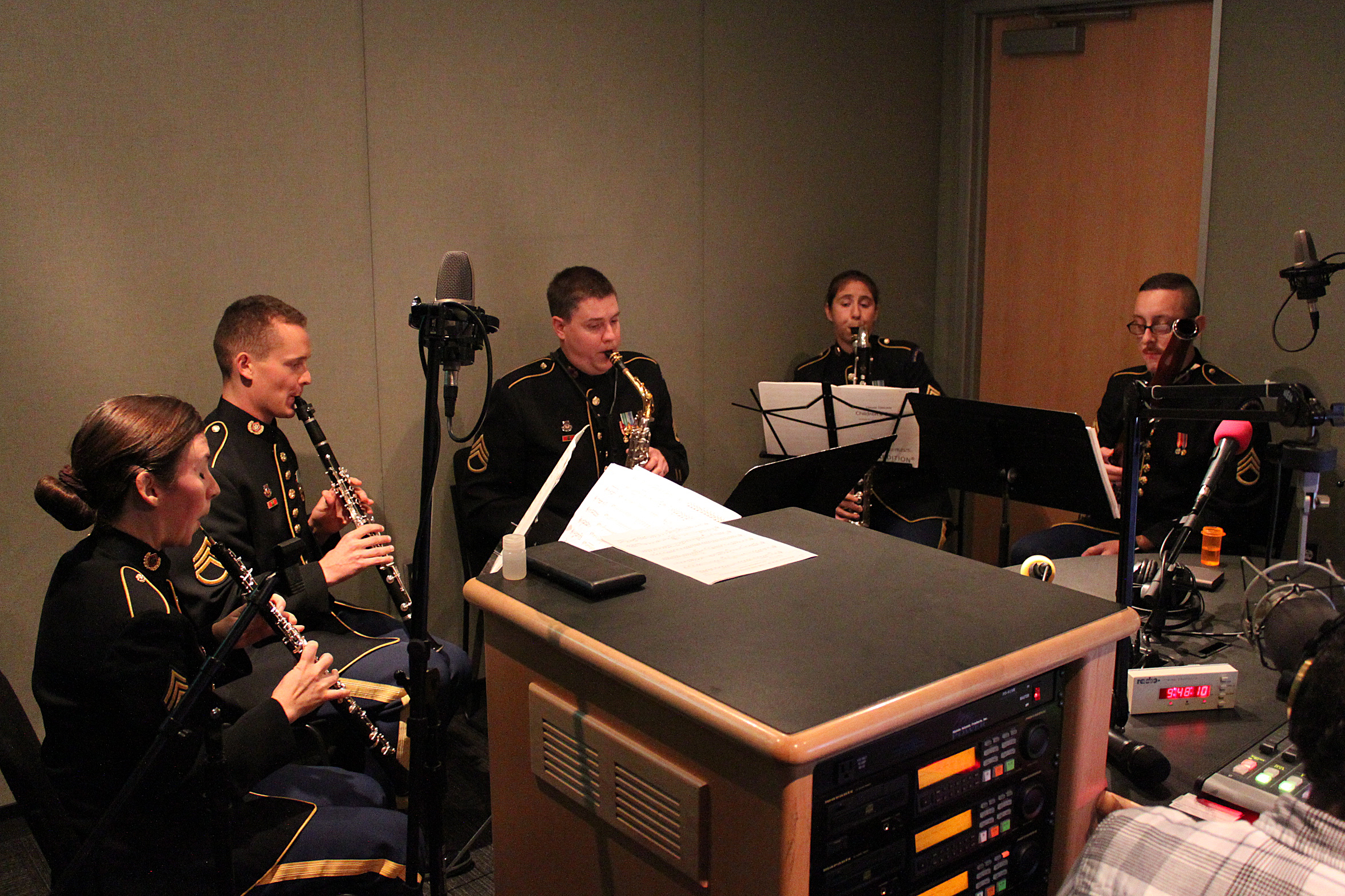 An adult playing oboe, an adult playing clarinet, an adult playing alto saxophone, an adult playing bass clarinet, and an adult playing bassoon, all looking at sheet music on music stands in a radio studio