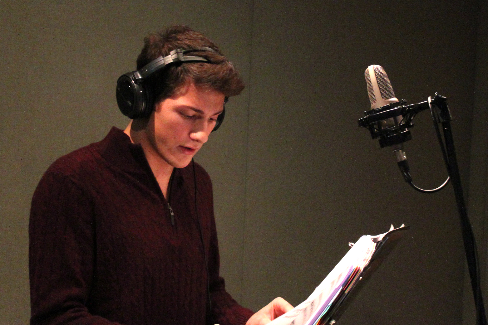 A teen wearing headphones looking down at paper in a binder standing behind a microphone in a radio studio