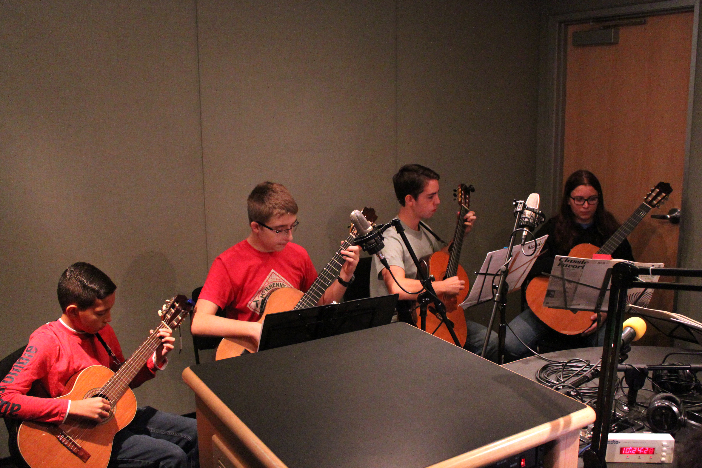 Four youth playing acoustic guitars looking at sheet music on music stands in a radio studio