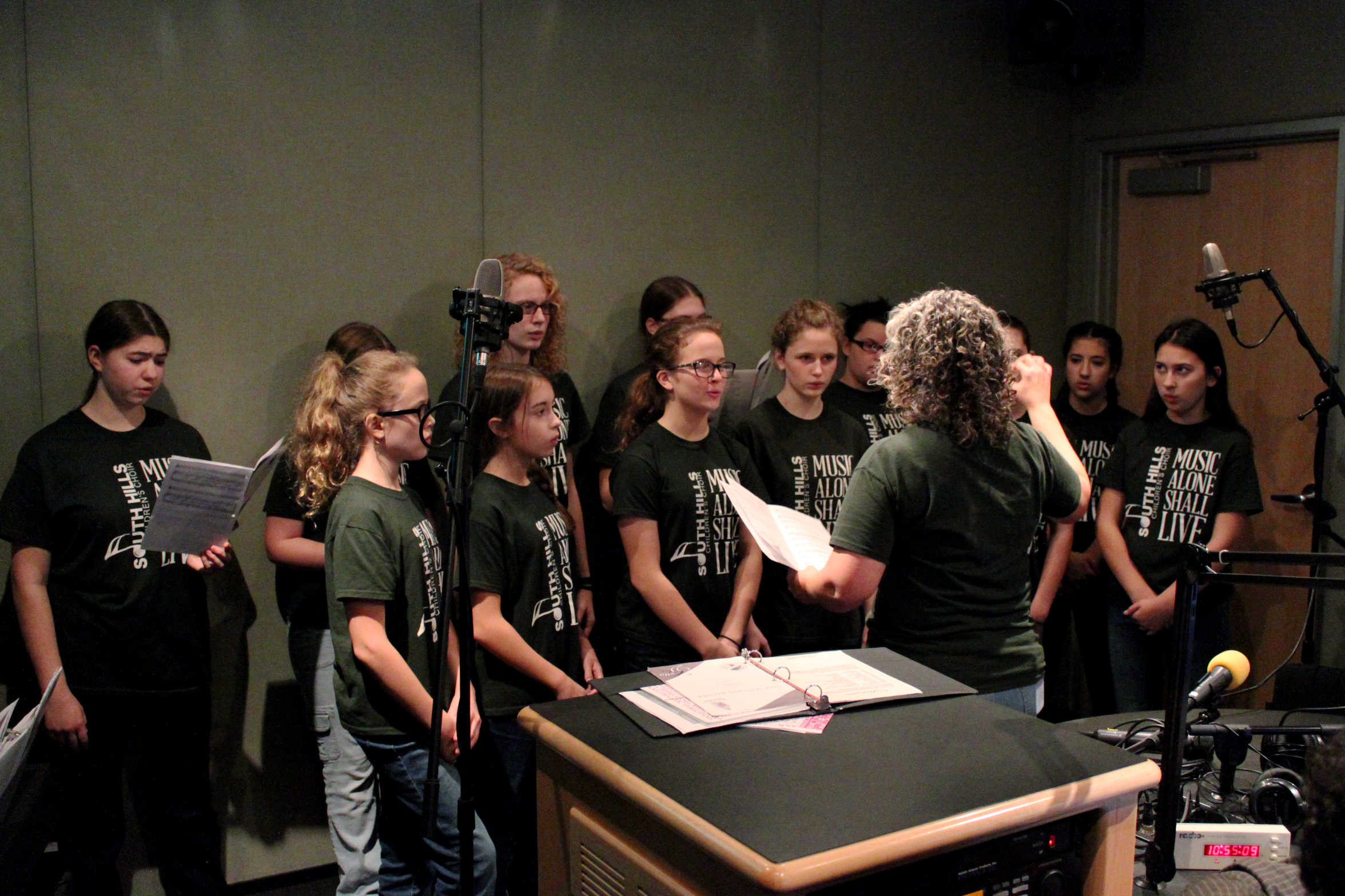 Twelve youth singing in a choir being led by an adult in a radio studio