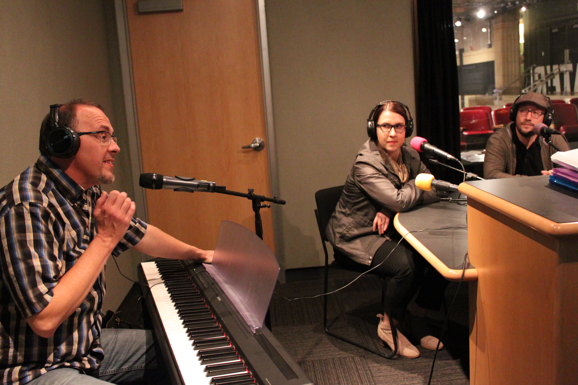 Three adults wearing headphones sitting behind microphones in a radio studio, one sitting behind a piano keyboard