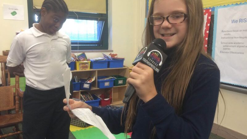 A youth looking at another youth smiling while holding a microphone and a piece of paper in a classroom