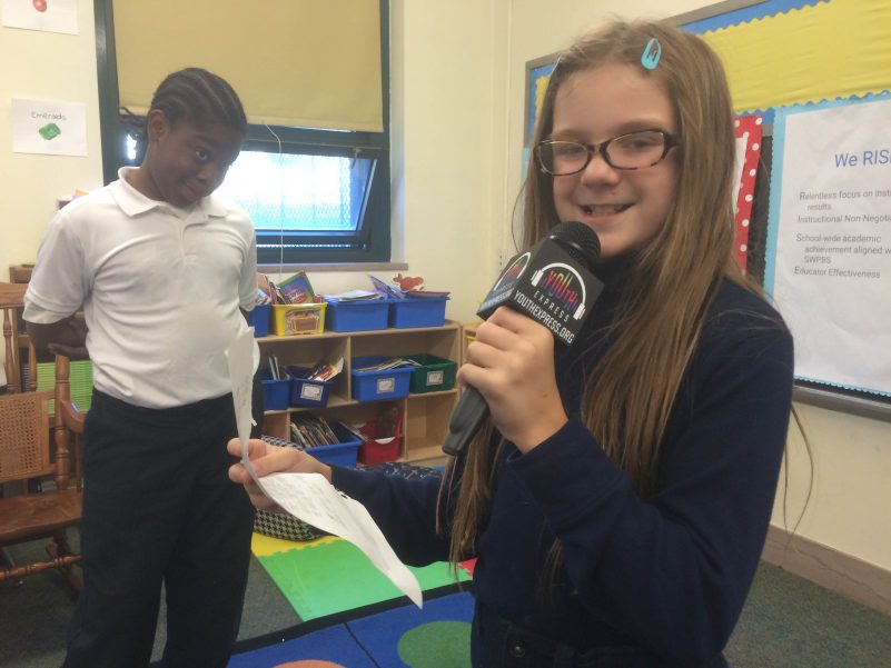 A youth looking at another youth smiling while holding a microphone and a piece of paper in a classroom