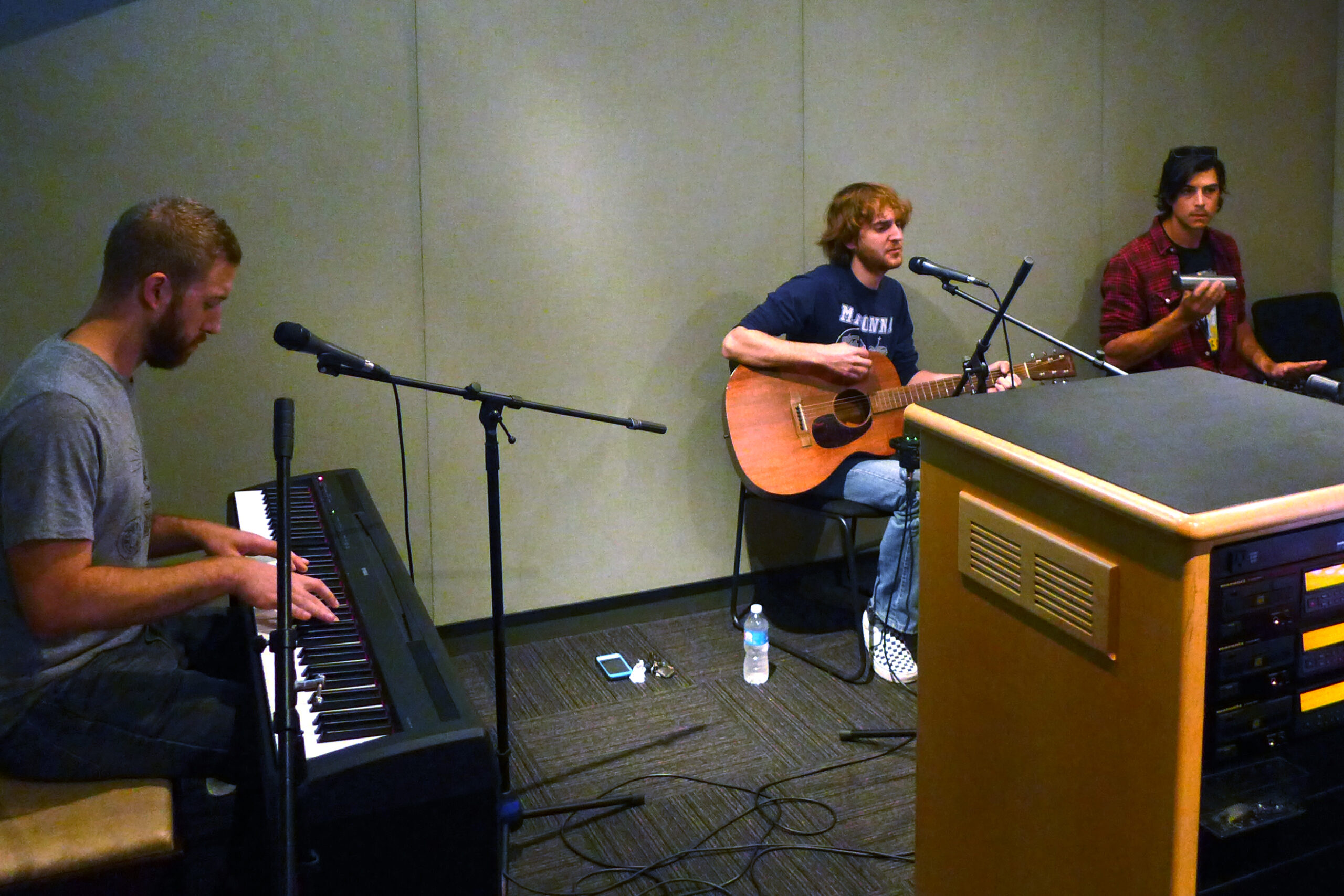 An adult playing a piano keyboard, an adult playing an acoustic guitar singing into a microphone, and an adult playing a shaker in a radio studio