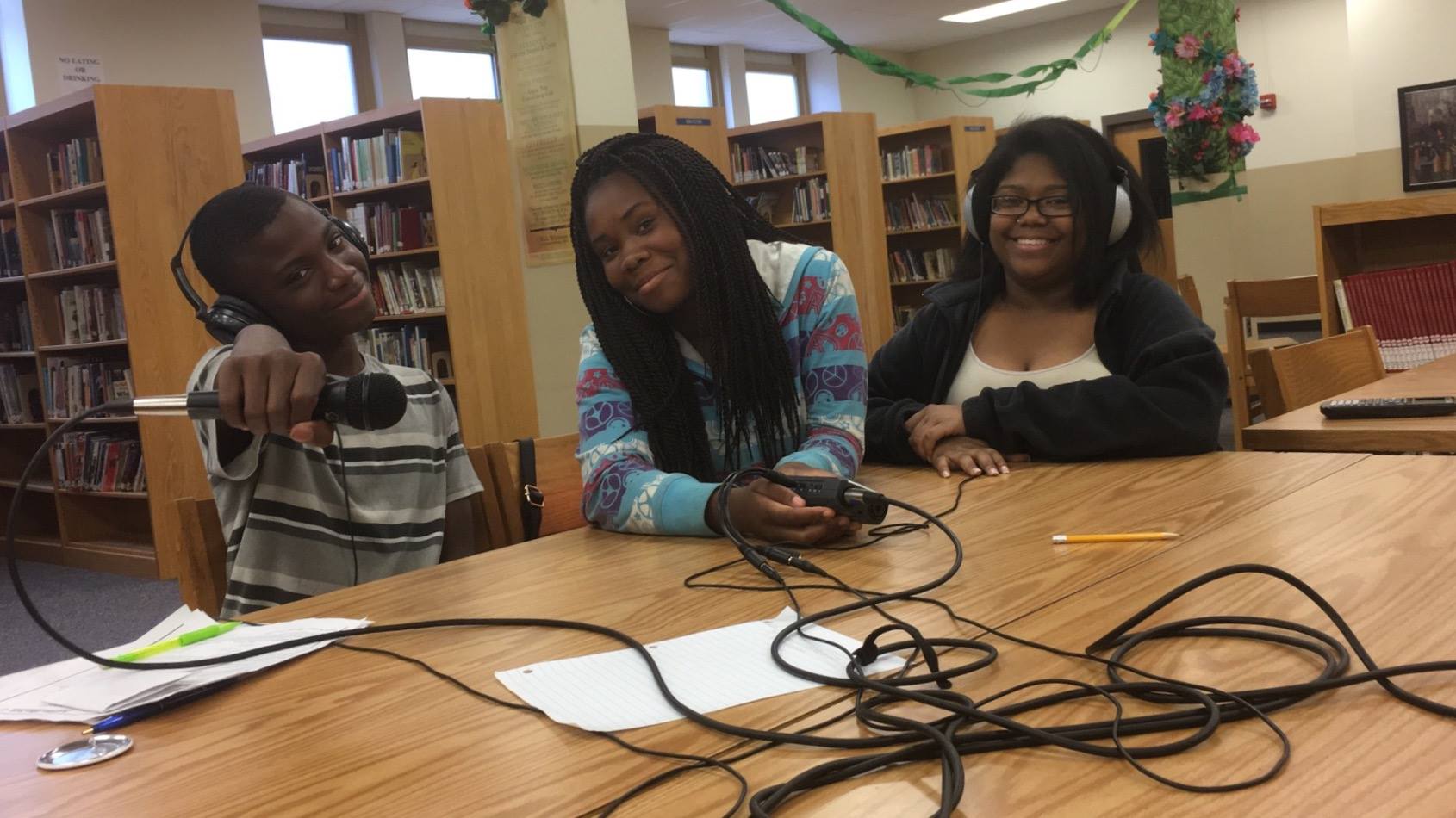 Three teens wearing headphones, one holding a microphone, one holding a portable recorder, sitting behind a wooden table in a library