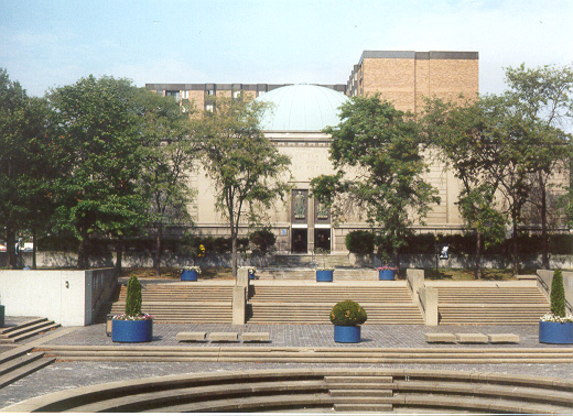 A wide shot of a large building with a dome on top, with a large set of stairs and five small shrubs in blue planters in front of the building