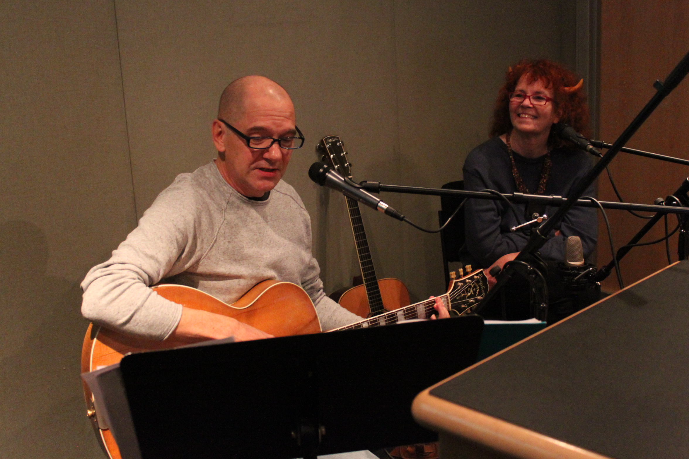 An adult singing into a microphone and playing an acoustic guitar and an adult smiling behind a microphone in a radio studio