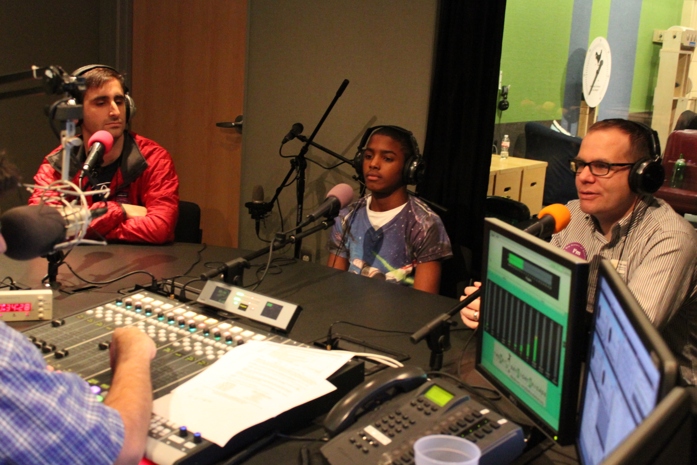 Two adults and a youth wearing headphones speaking into microphones being interviewed in a radio studio