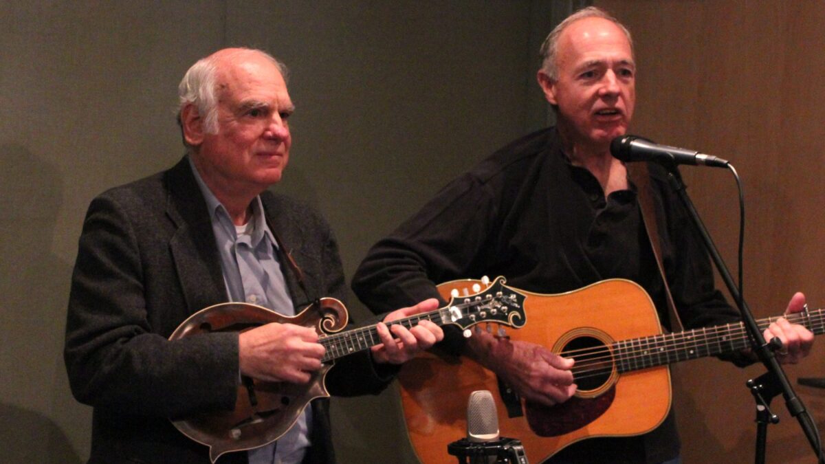 An adult playing a mandolin and an adult playing an acoustic guitar and singing into a microphone in a radio studio