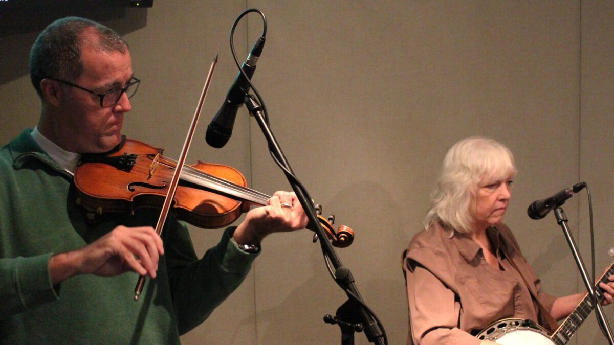 An adult playing a fiddle into a microphone and an adult playing a banjo and singing into a microphone in a radio studio