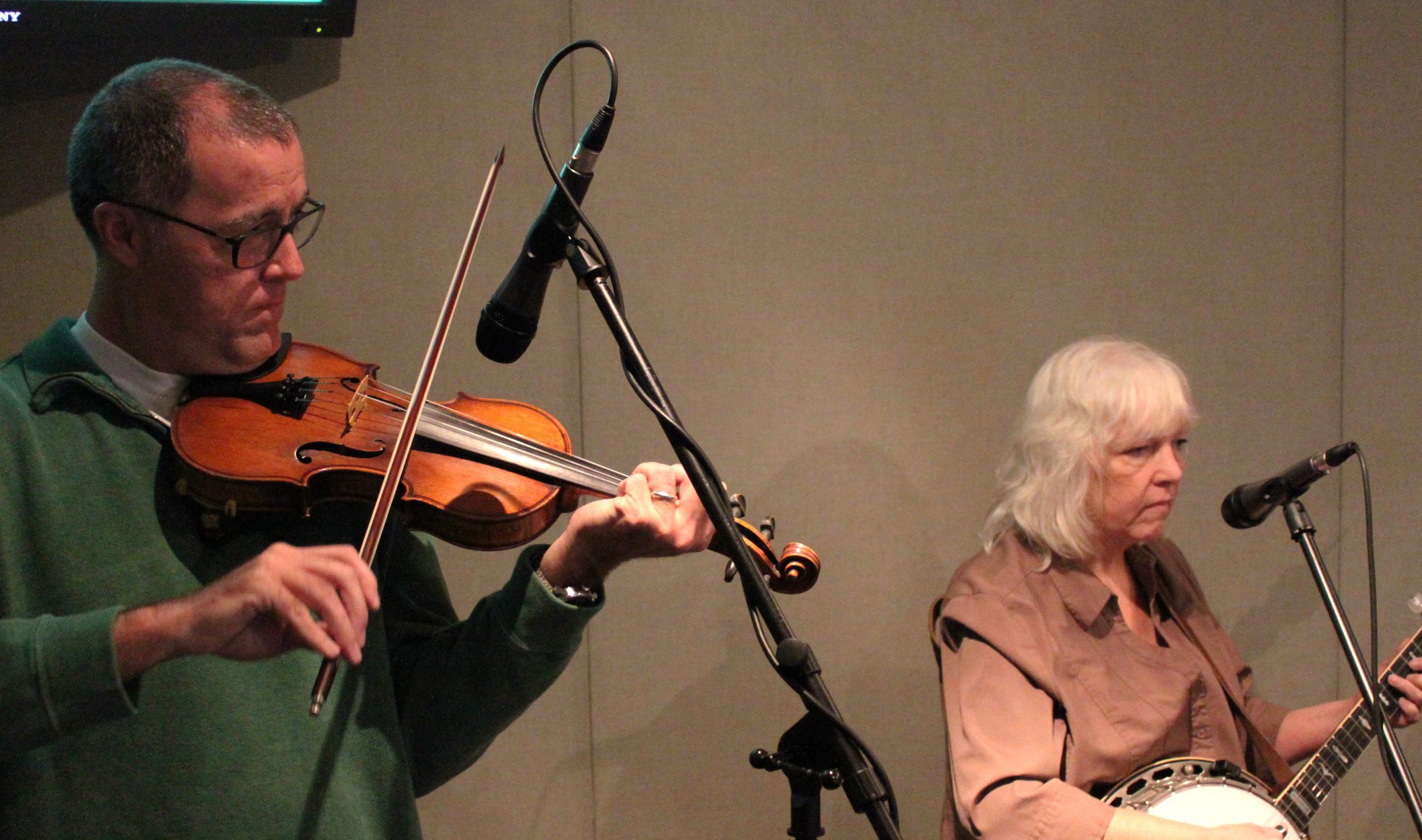 An adult playing a fiddle into a microphone and an adult playing a banjo and singing into a microphone in a radio studio