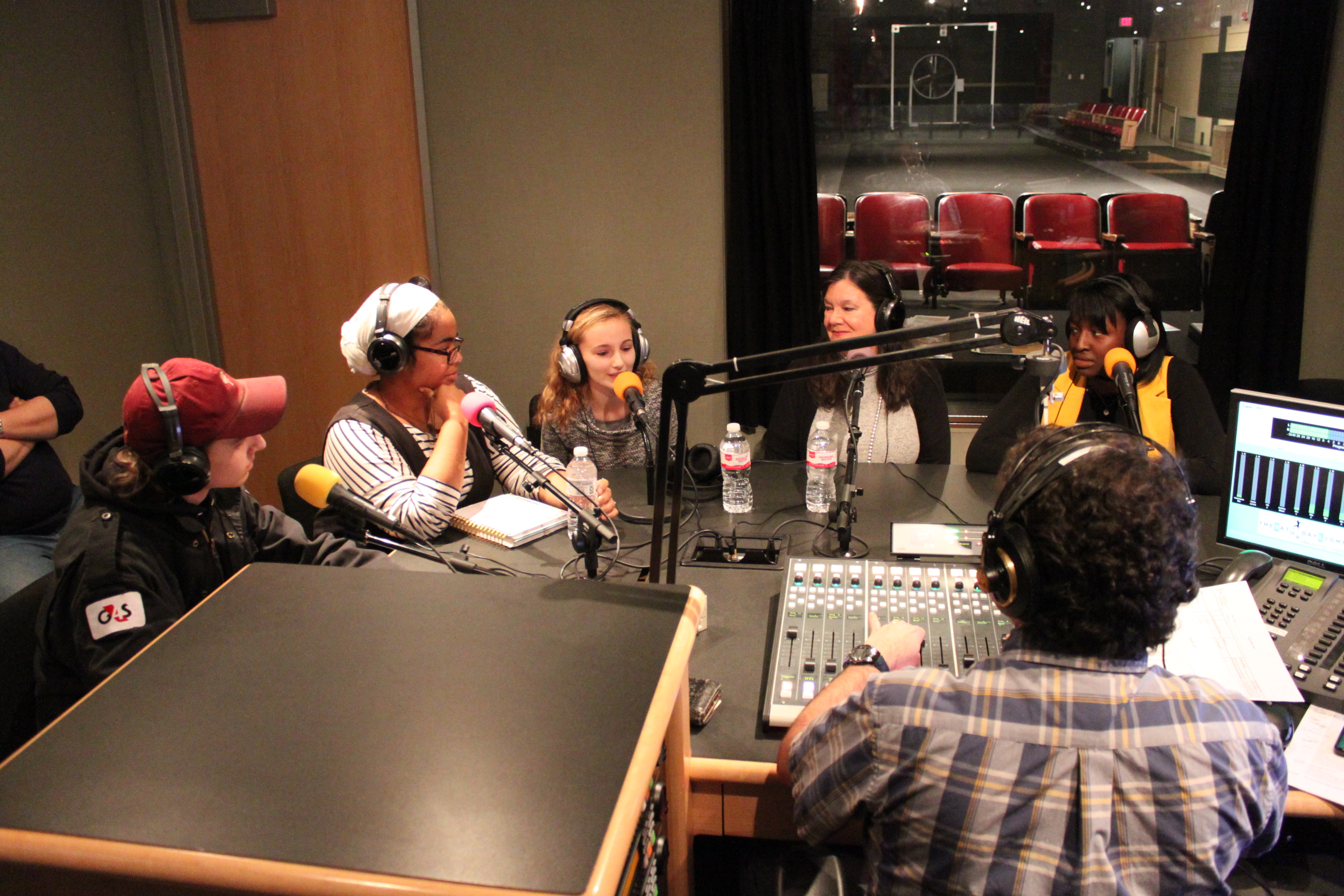 Five teens wearing headphones speaking into microphones being interviewed by an adult wearing headphones sitting behind a microphone and a mixing board in a radio studio