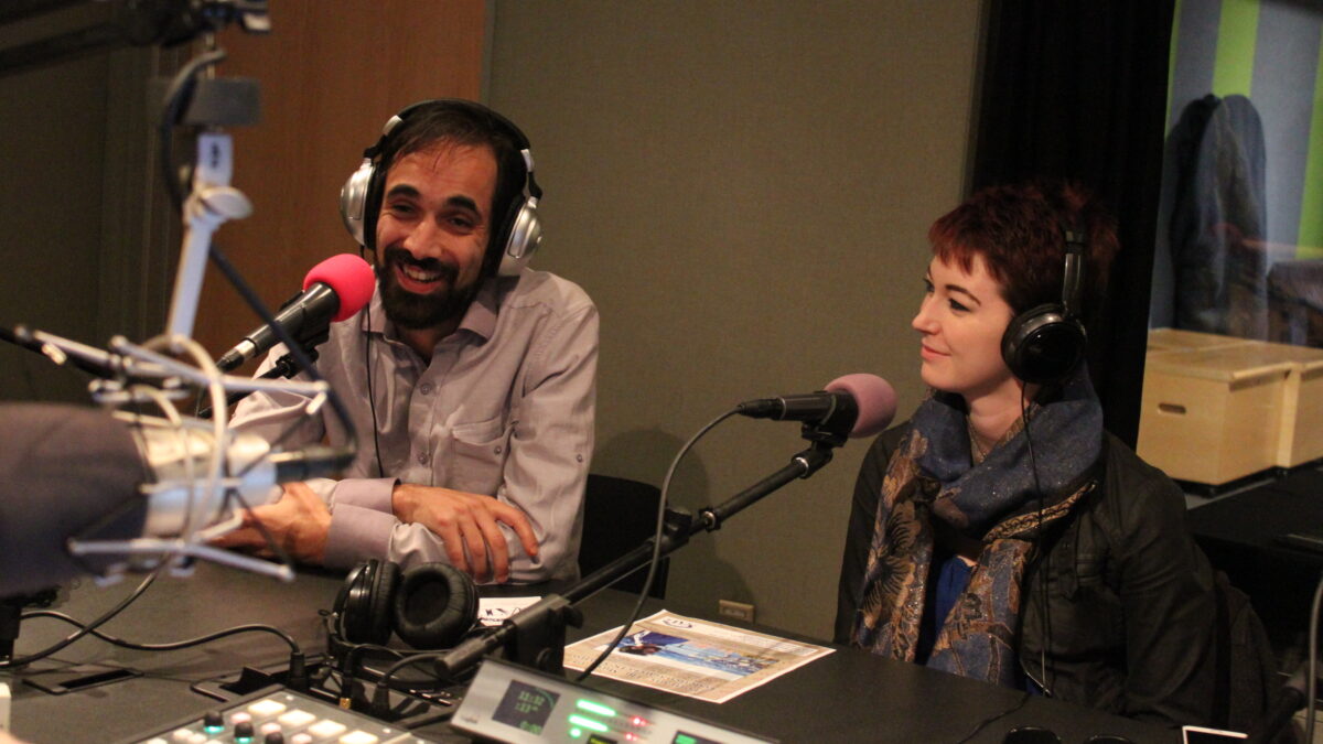 Two adults wearing headphones speaking into microphones in a radio studio