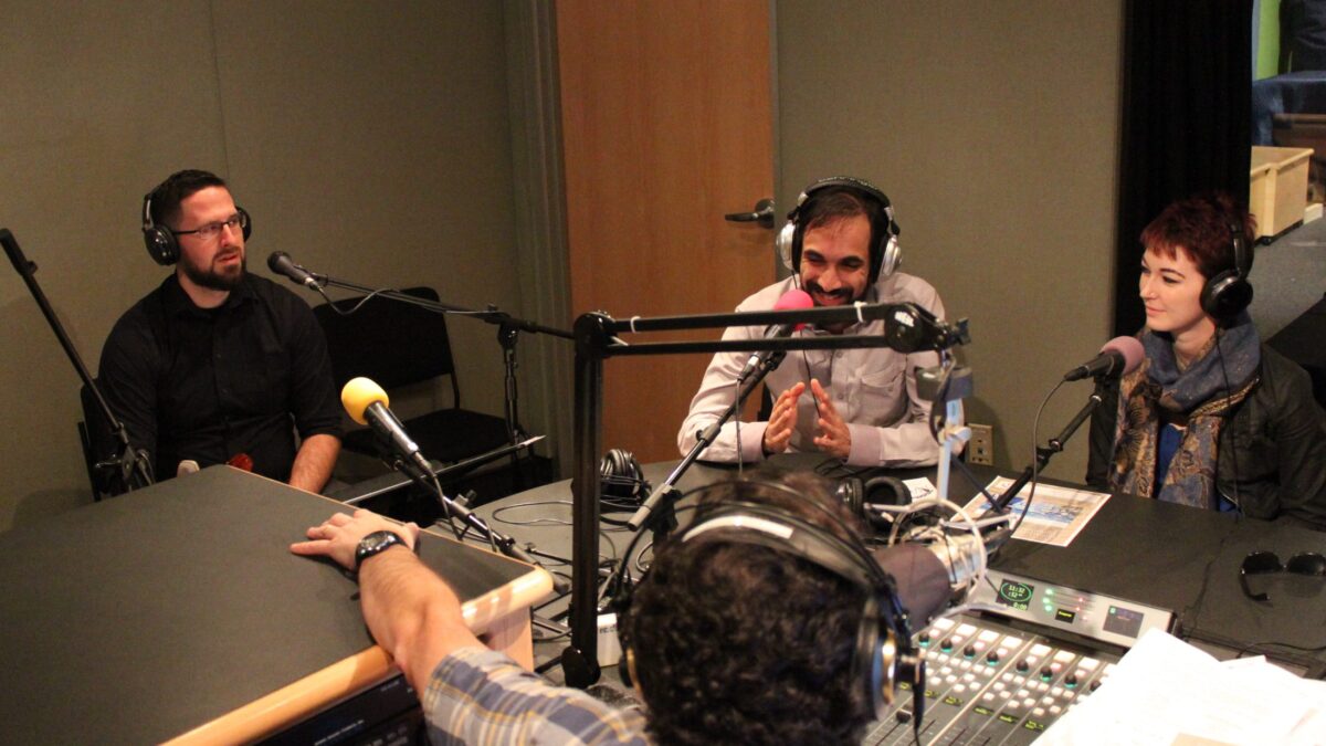 Three adults wearing headphones speaking into microphones being interviewed by an adult wearing headphones sitting behind a microphone and a mixing board in a radio studio