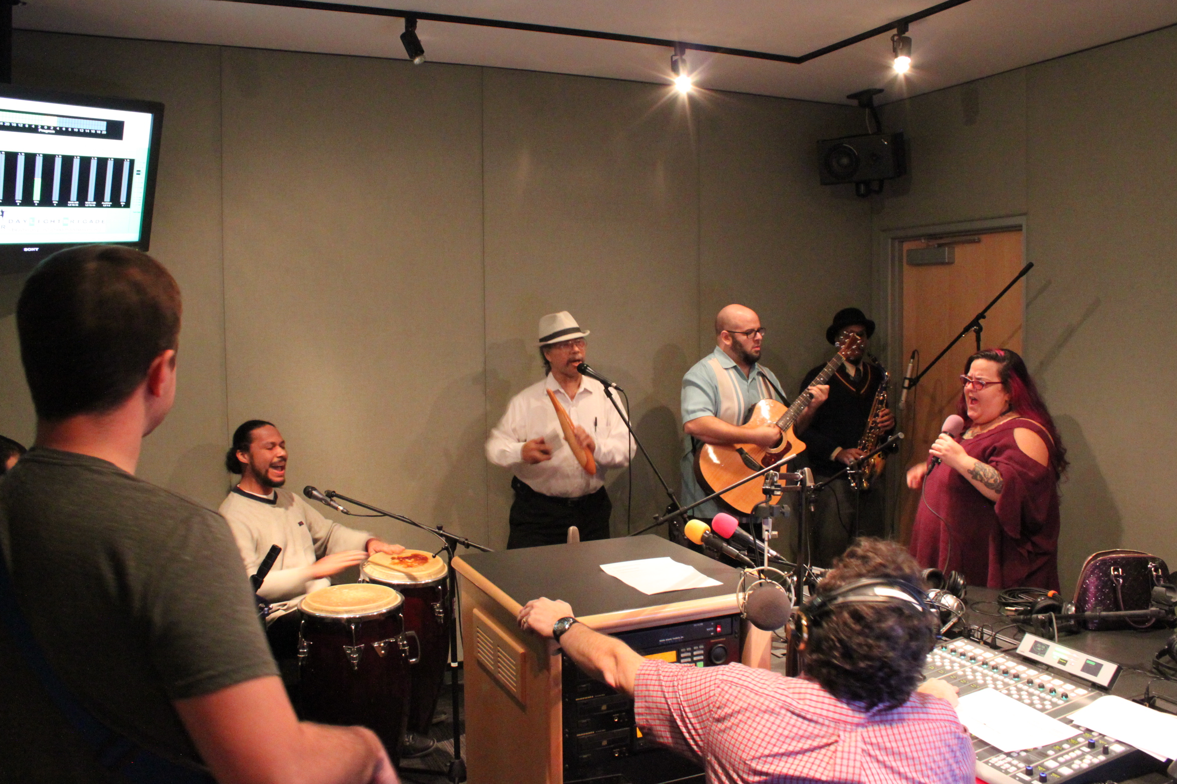 An adult singing into a microphone, an adult playing an acoustic guitar, two adults playing percussive instruments, and an adult holding an instrument as another adult wearing headphones sitting behind a microphone and a mixing board listens in a radio studio