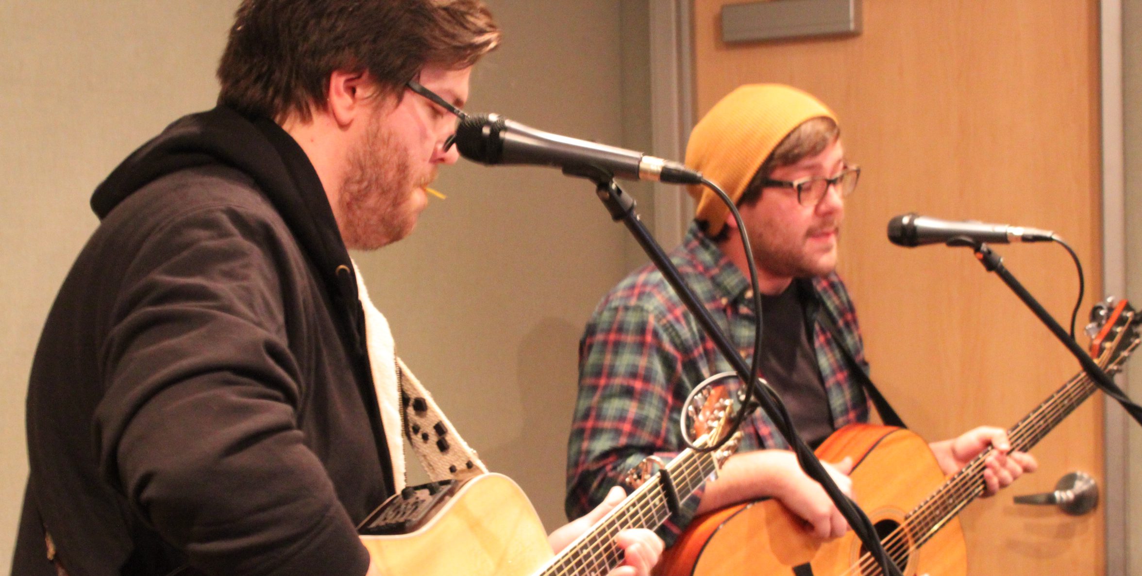 Two adults playing acoustic guitars singing into microphones in a radio studio