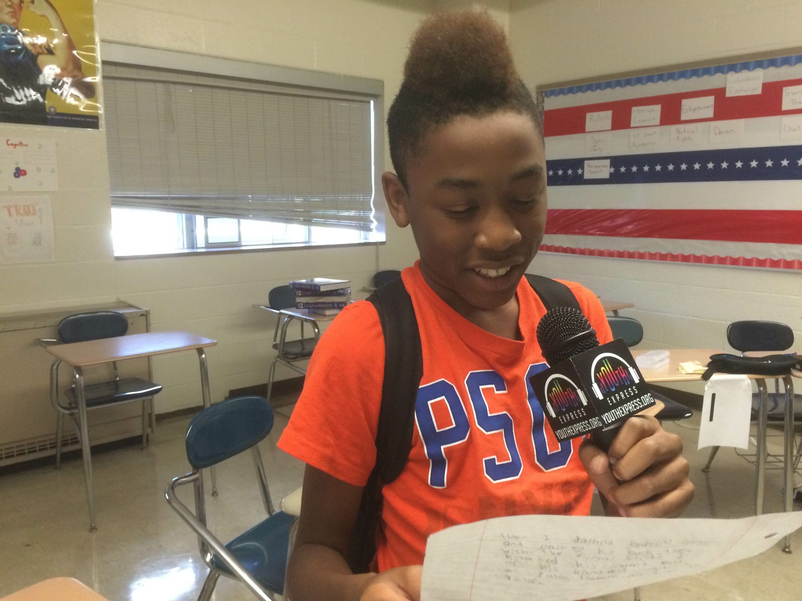 A youth holding a piece of paper and speaking into a microphone in a classroom