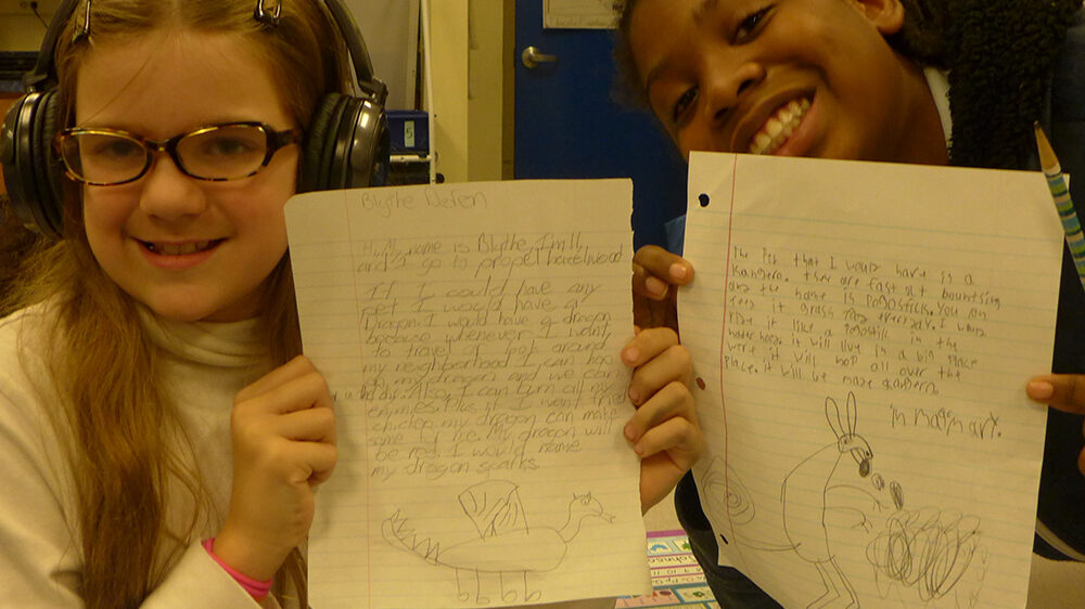 Two youth wearing headphones holding pieces of notebook paper up to the camera in a classroom