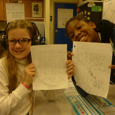 Two youth wearing headphones holding pieces of notebook paper up to the camera in a classroom