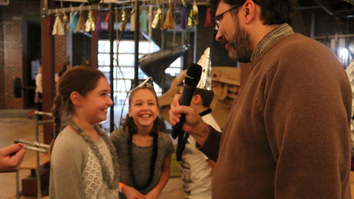 An adult holding and speaking into a microphone interviewing two smiling youth