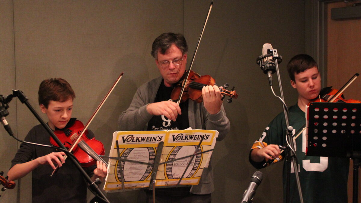 Two youth and one adult playing violins looking at sheet music on music stands in a radio studio