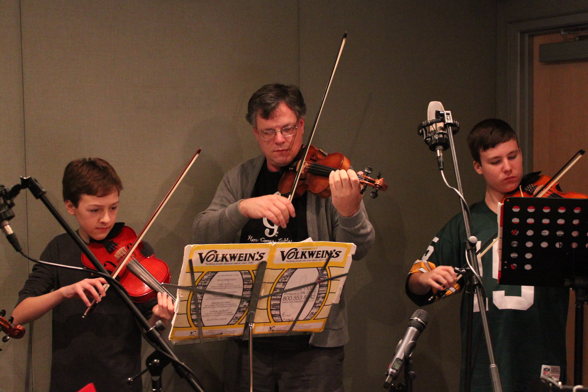 Two youth and one adult playing violins looking at sheet music on music stands in a radio studio
