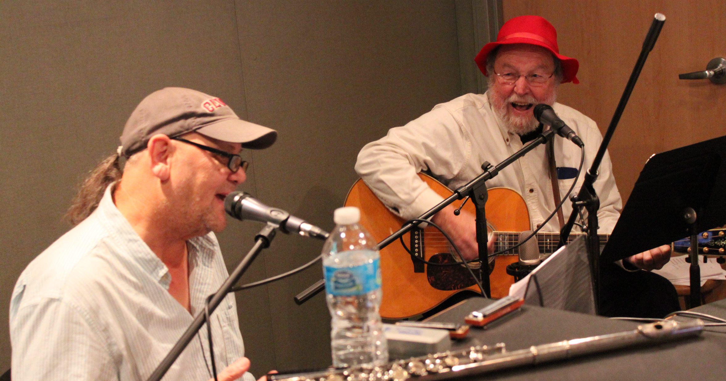 Two adults singing into microphones in a radio studio, one playing an acoustic guitar