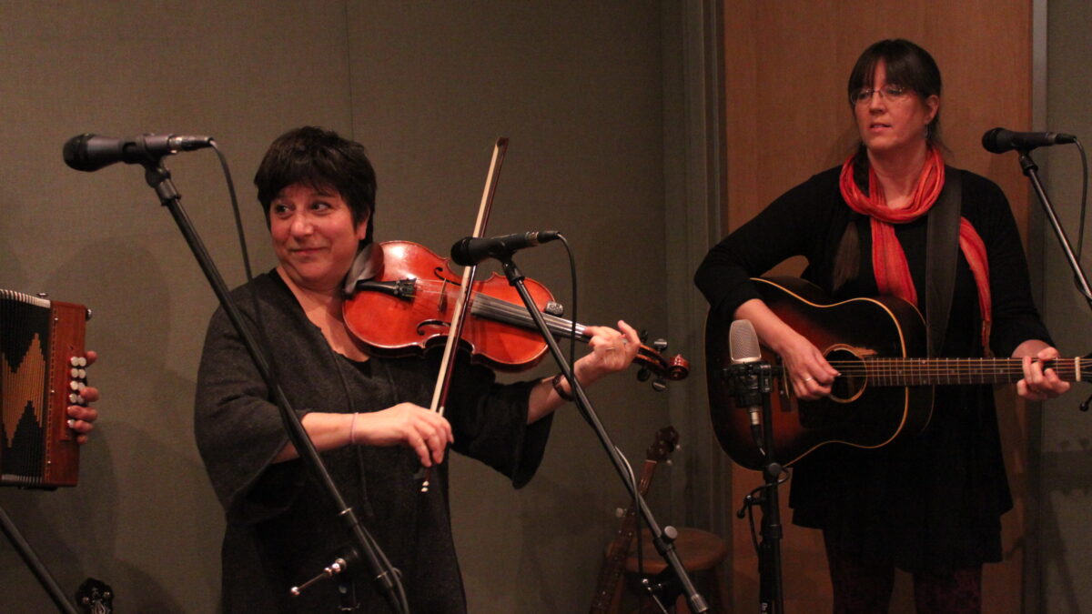 An adult playing fiddle and an adult playing an acoustic guitar in a radio studio