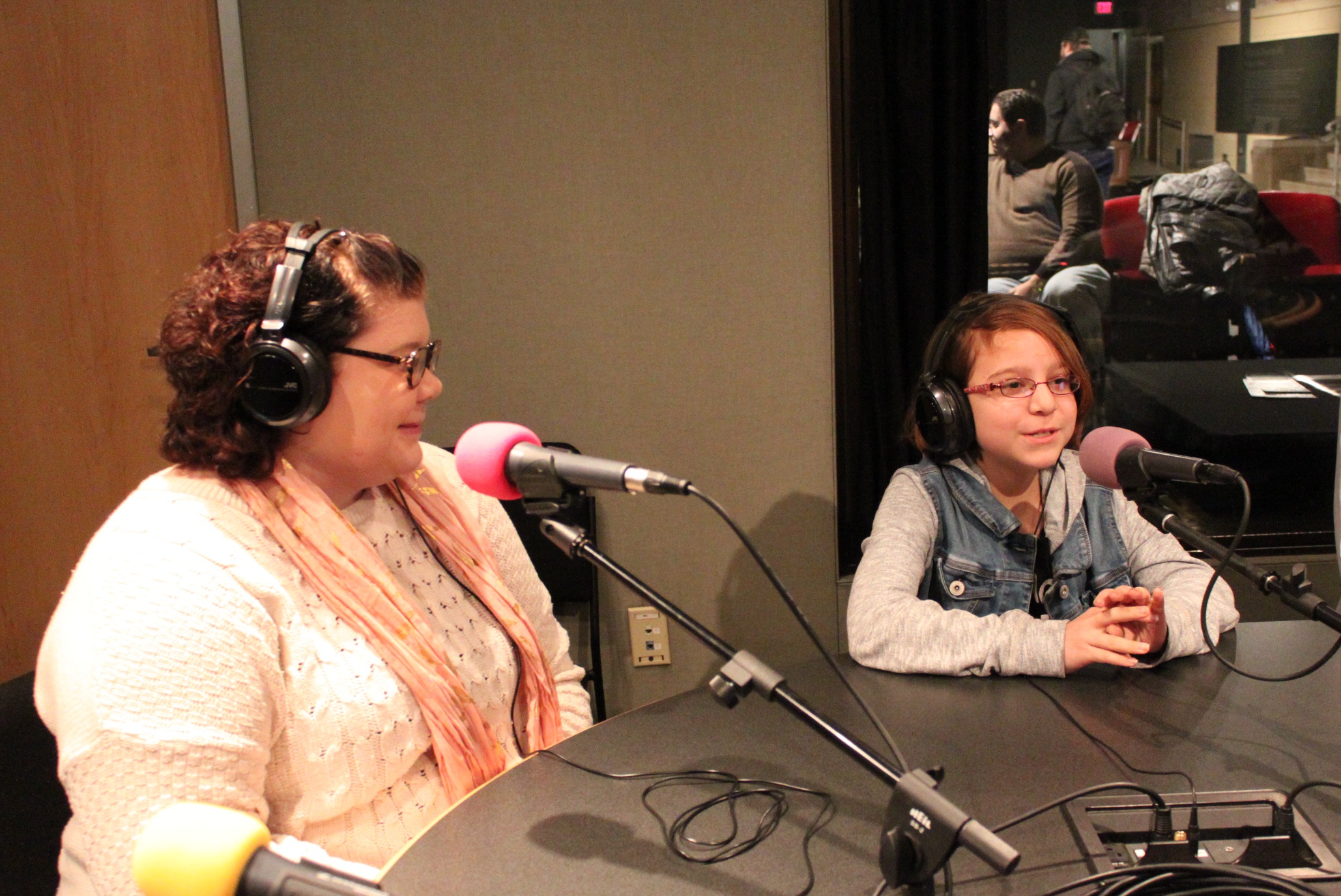An adult and a youth wearing headphones speaking into microphones in a radio studio