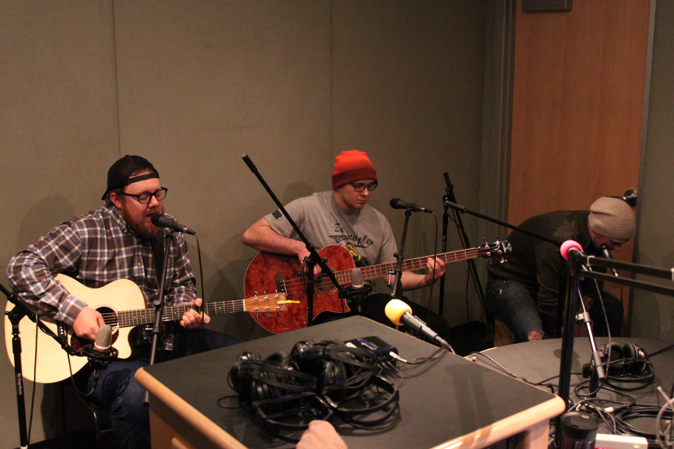 An adult playing acoustic guitar and singing into microphones, an adult playing electric bass, and an adult playing a cajon in a radio studio