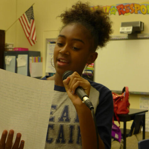 A youth holding a piece of paper reading into a microphone in a classroom