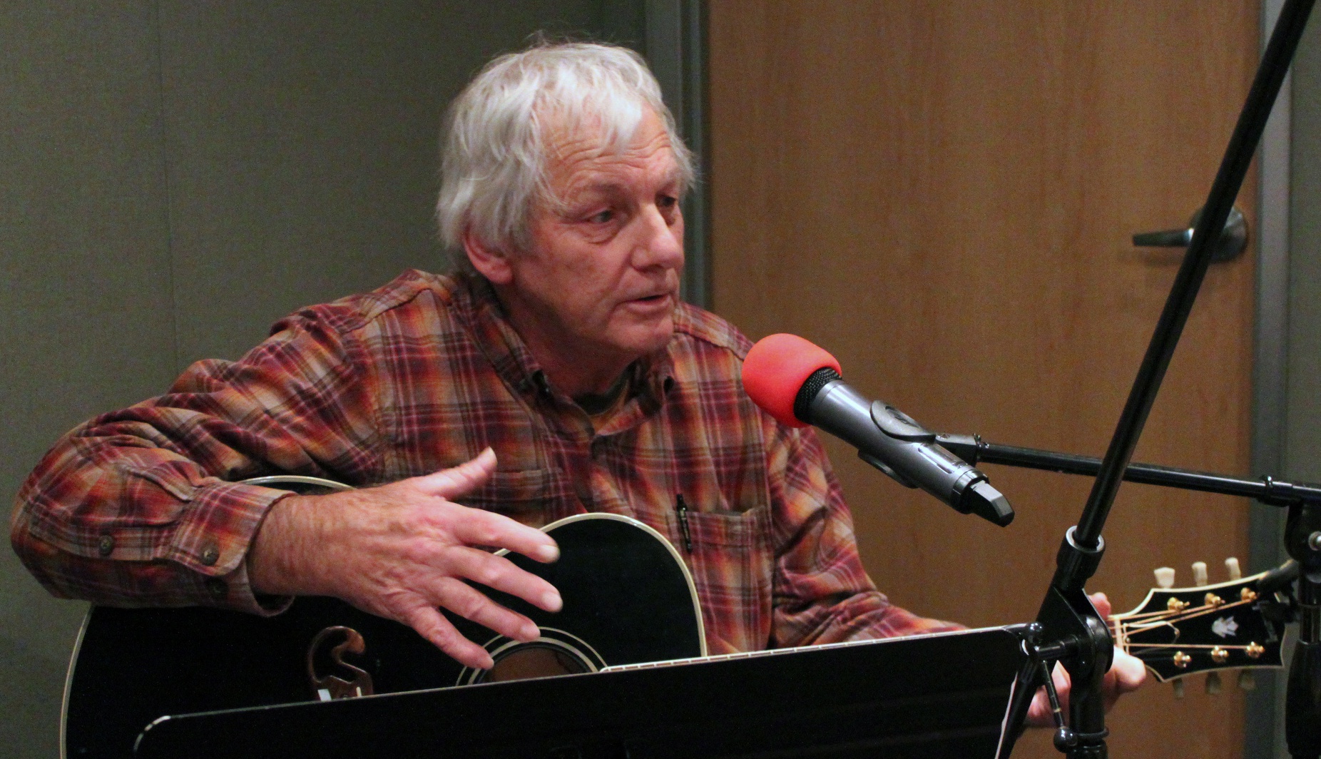 An adult holding an acoustic guitar and singing into a microphone in a radio studio