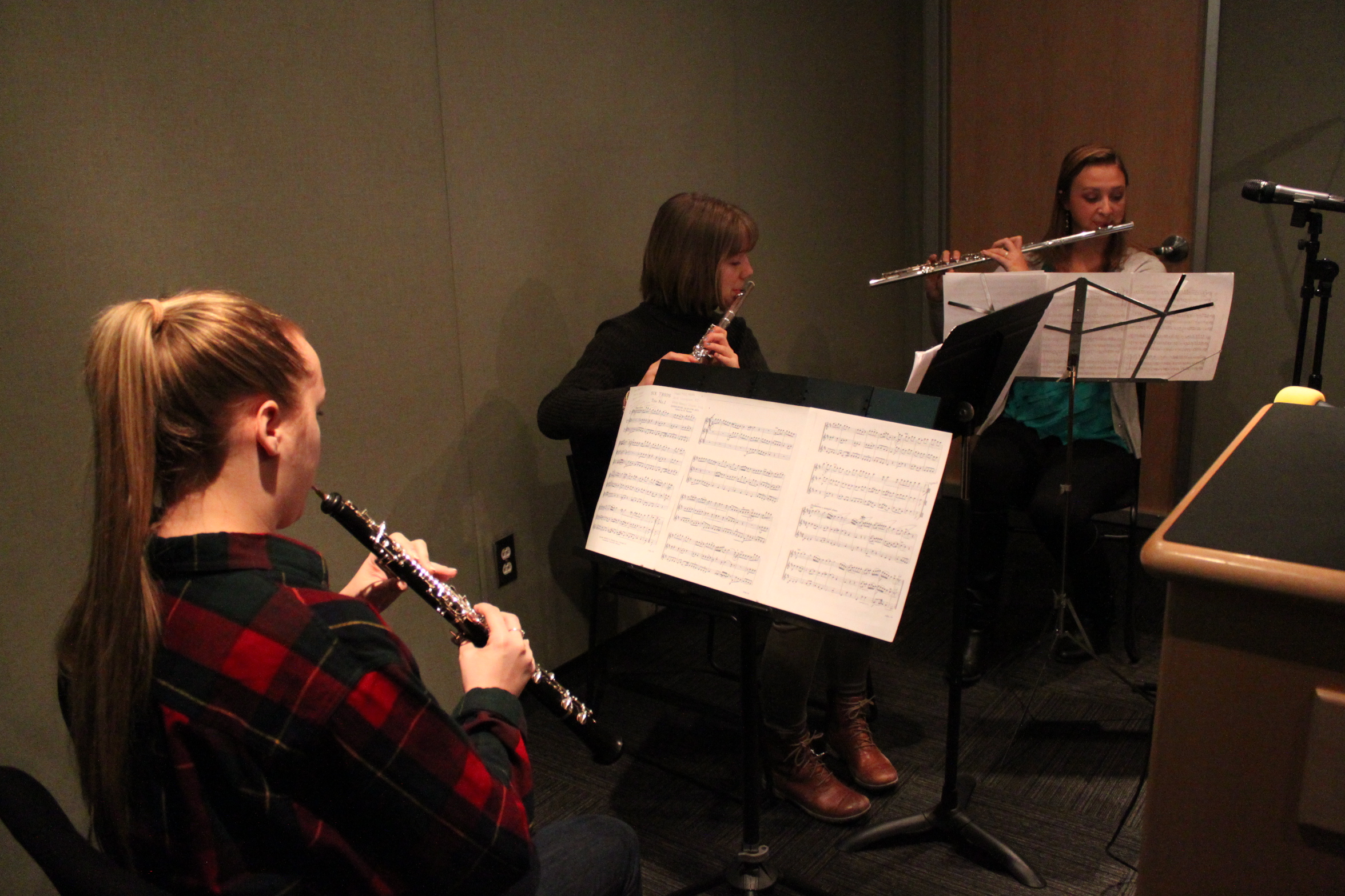 Two teens playing flute and one teen playing oboe looking at sheet music on music stands in a radio studio
