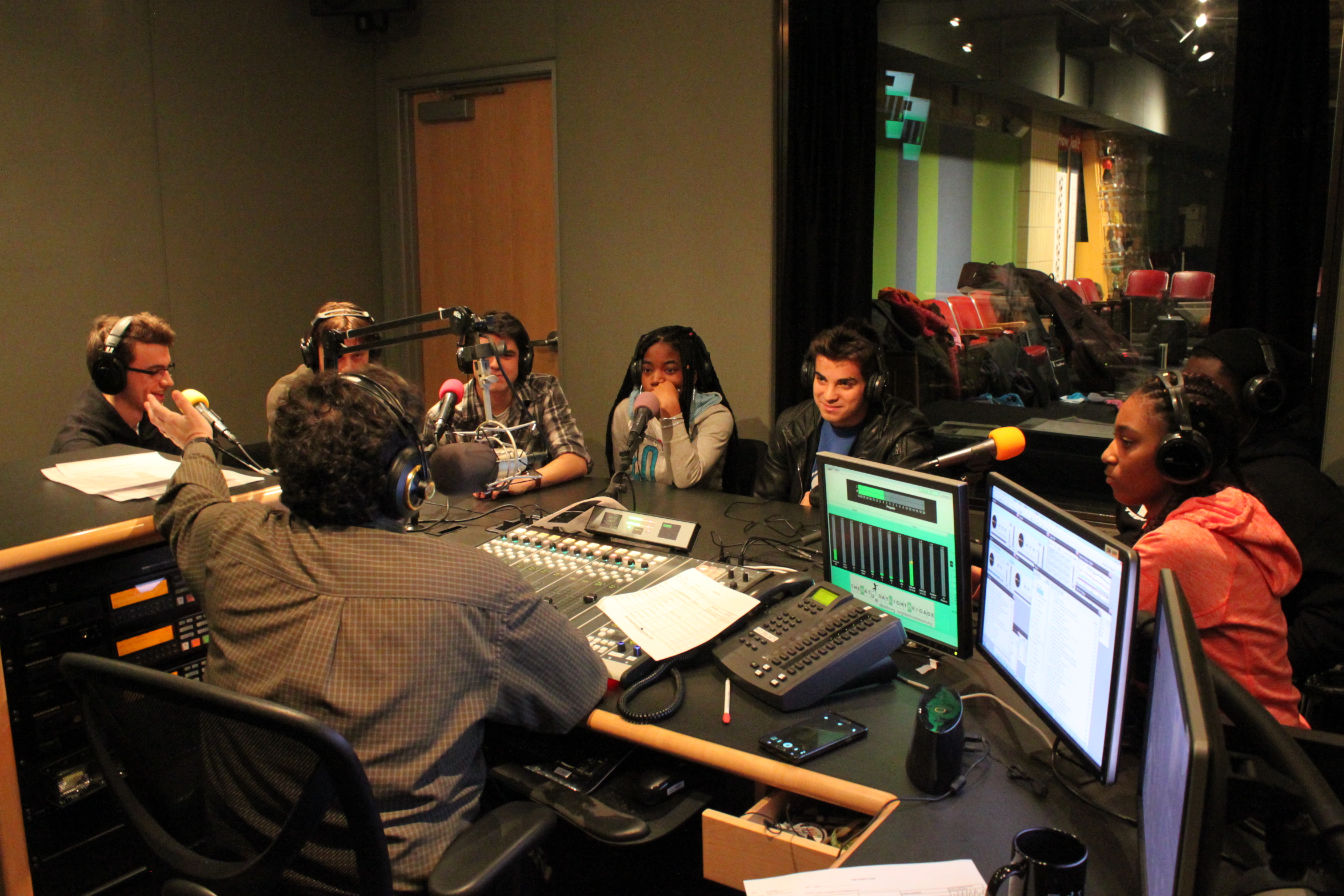 Six teens wearing headphones speaking into microphones being interviewed by an adult wearing headphones sitting behind a microphone and a mixing board in a radio studio.