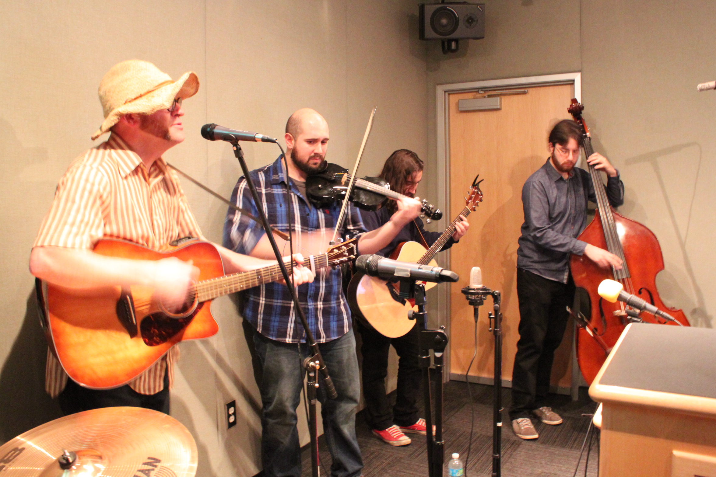 Two adults playing acoustic guitar, an adult playing fiddle, and an adult playing upright bass in a radio studio