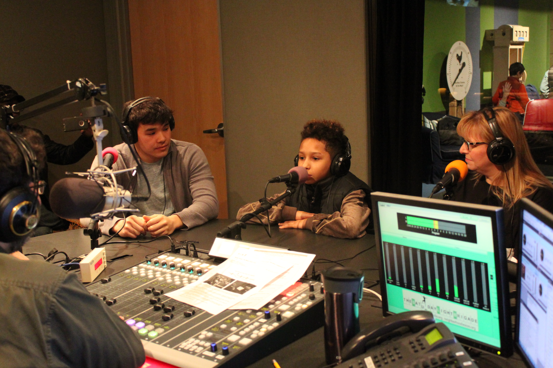 Two adults and a youth wearing headphones speaking into microphones being interviewed by an adult wearing headphones sitting behind a microphone and a mixing board in a radio studio