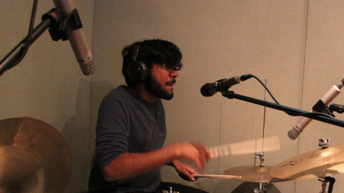 An adult wearing headphones playing a drum set in a radio studio