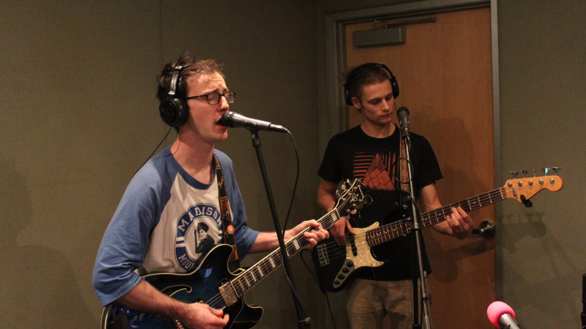 An adult wearing headphones playing electric guitar and singing into a microphone and an adult wearing headphones playing electric bass in a radio studio