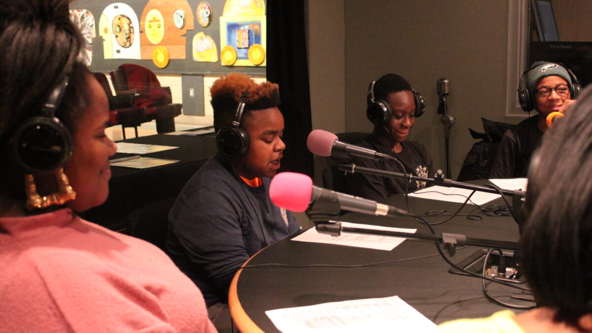 Five youth wearing headphones reading from pieces of paper into microphones in a radio studio