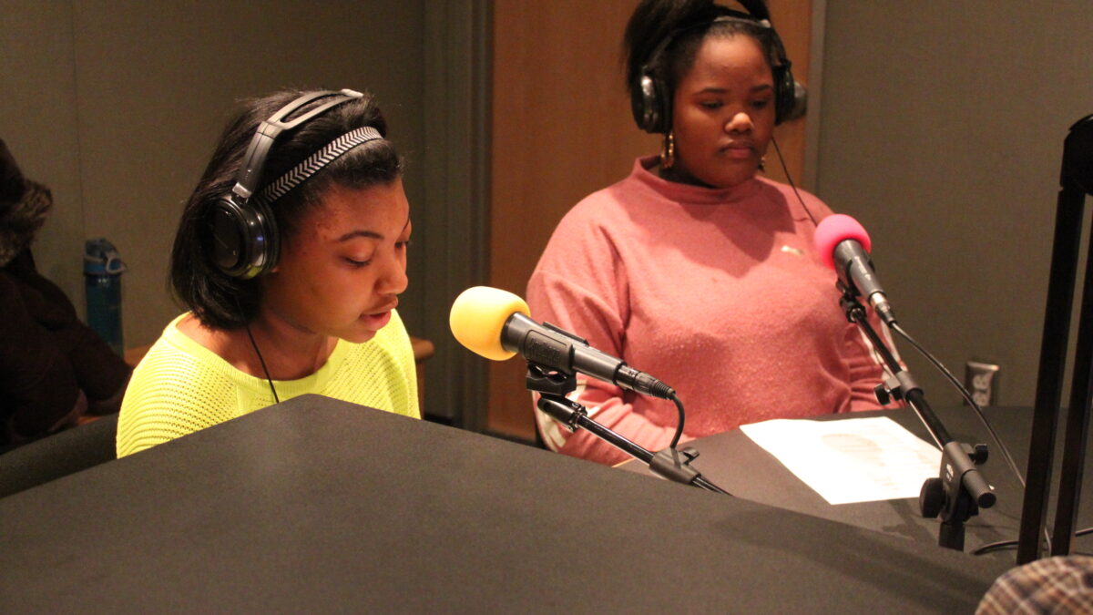 Two teens wearing headphones reading pieces of paper into microphones in a radio studio