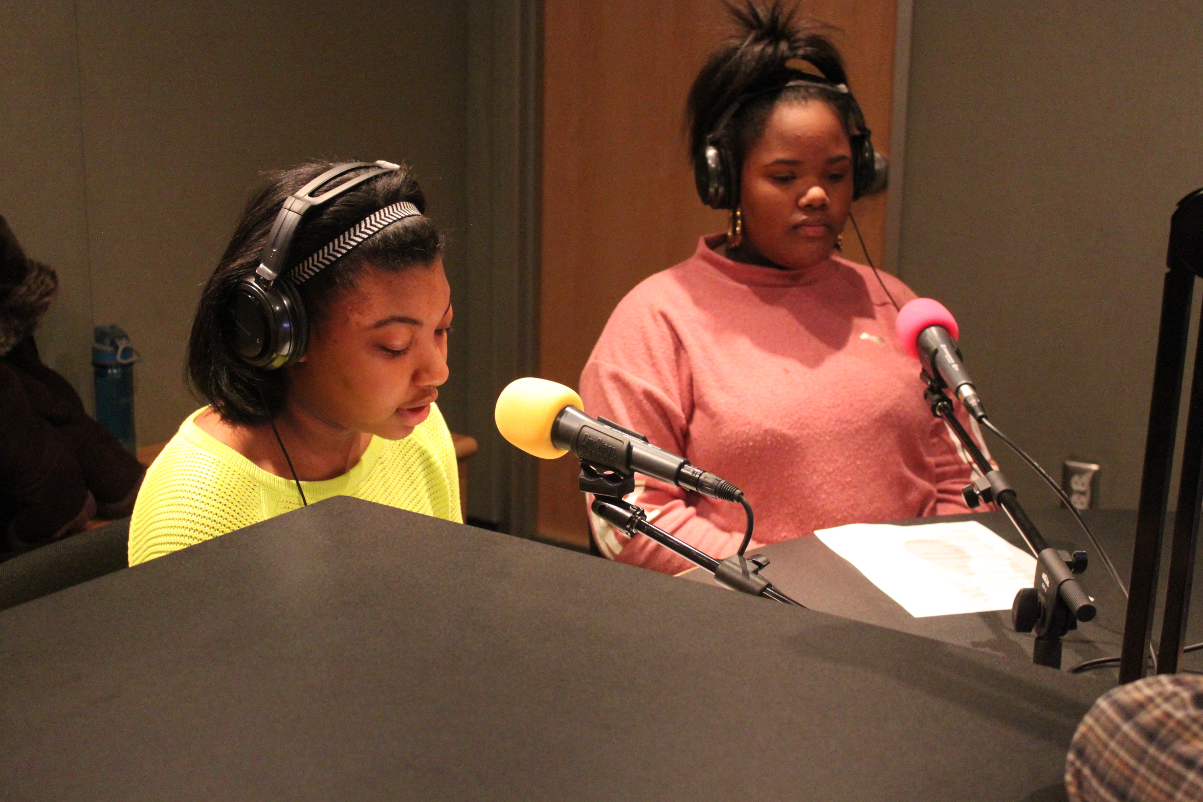 Two teens wearing headphones reading pieces of paper into microphones in a radio studio