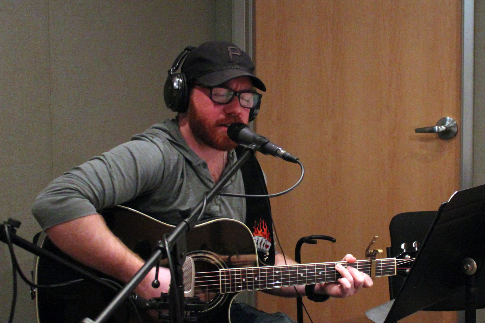 An adult wearing headphones singing into a microphone and playing acoustic guitar in a radio studio