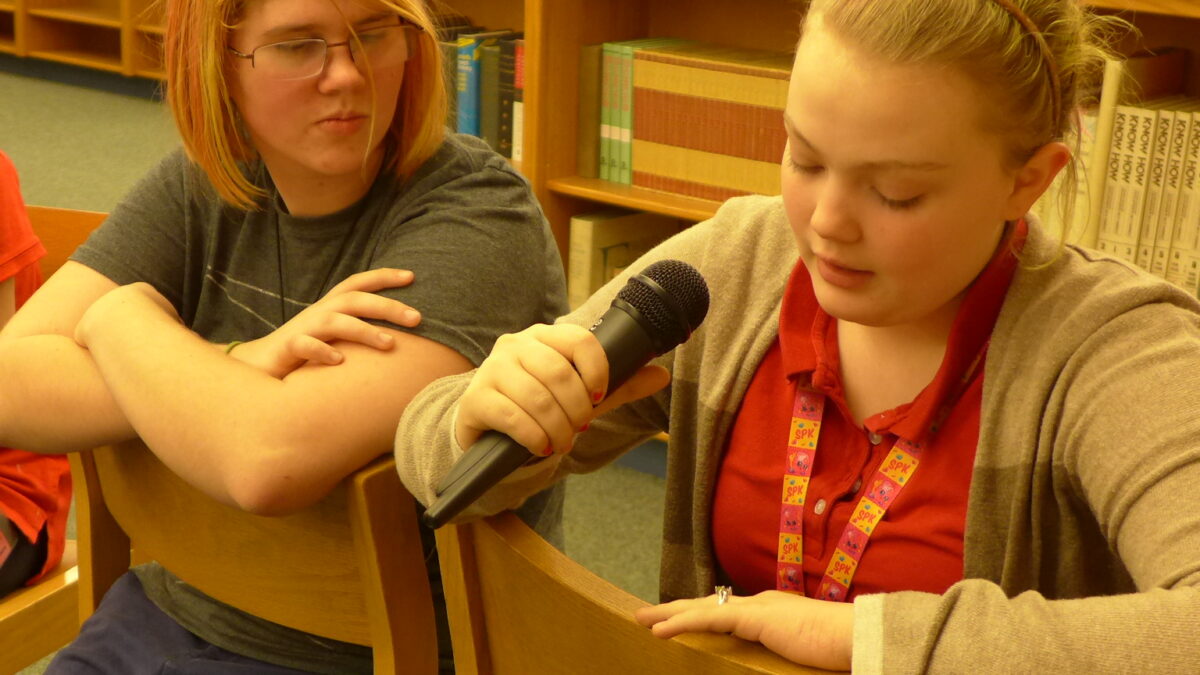 Two teens sitting in a library, one holding and speaking into a microphone