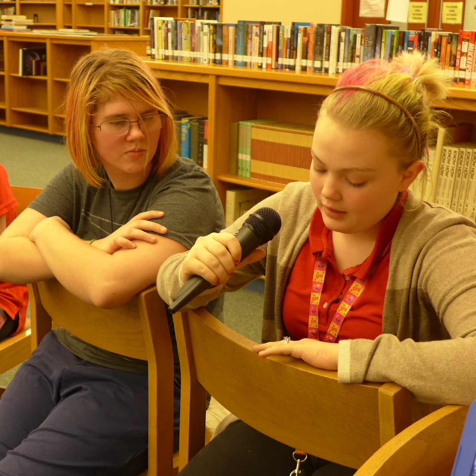 Two teens sitting in a library, one holding and speaking into a microphone