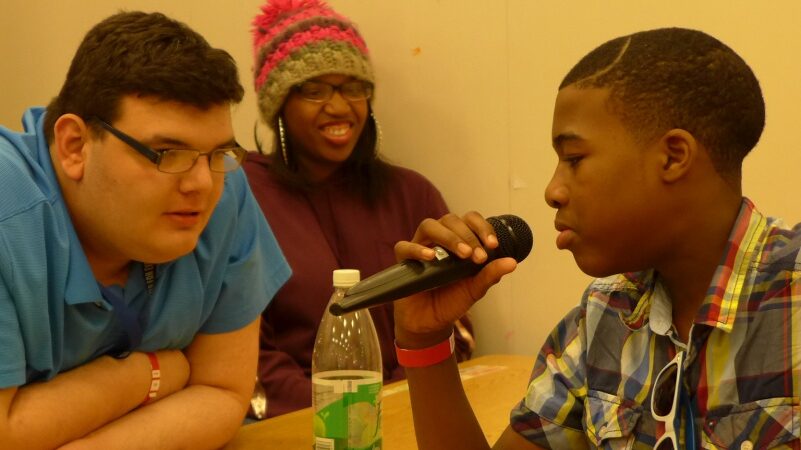 Three teens sitting around a table in a classroom, one speaking into a microphone