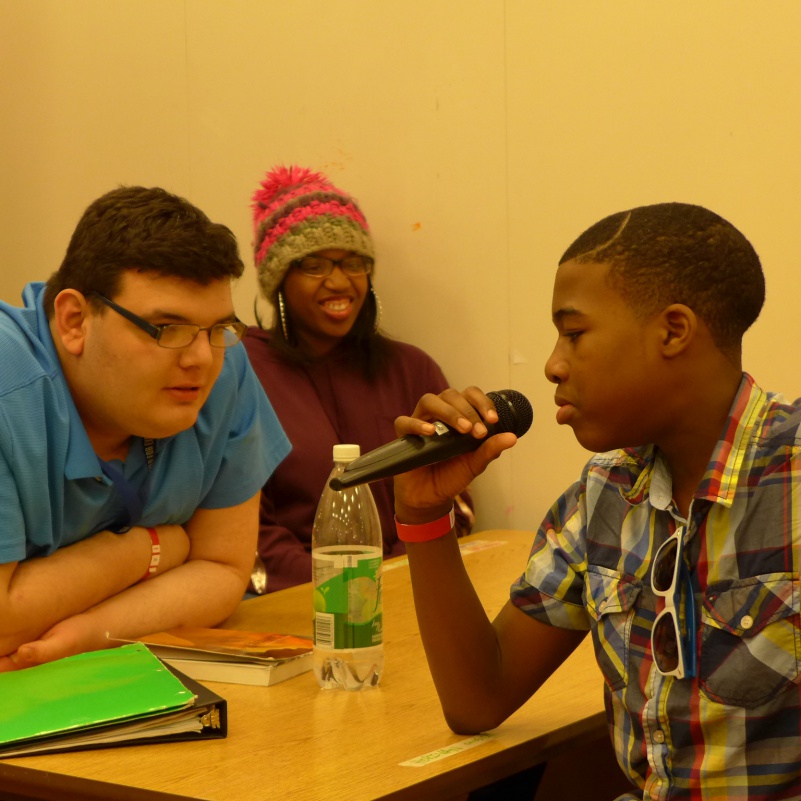 Three teens sitting around a table in a classroom, one speaking into a microphone