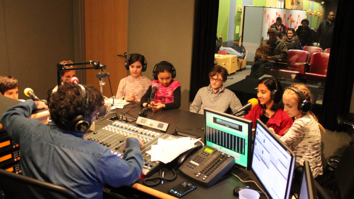 Seven youth wearing headphones sitting behind microphones being interviewed by an adult wearing headphones sitting behind a microphone and a mixing board in a radio studio