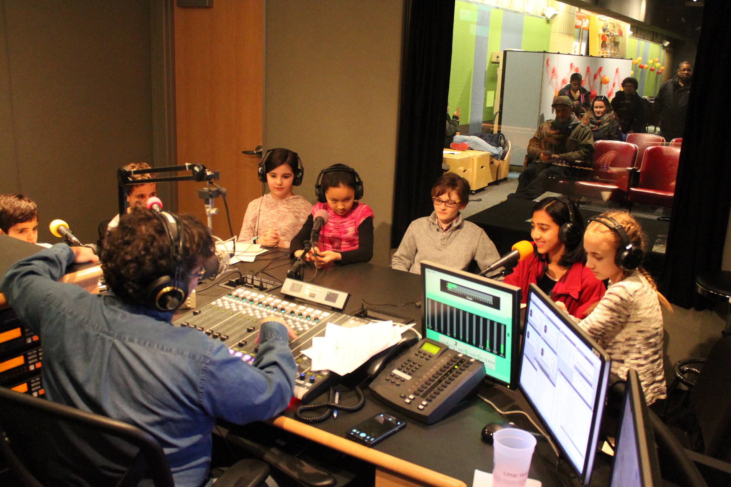 Seven youth wearing headphones sitting behind microphones being interviewed by an adult wearing headphones sitting behind a microphone and a mixing board in a radio studio